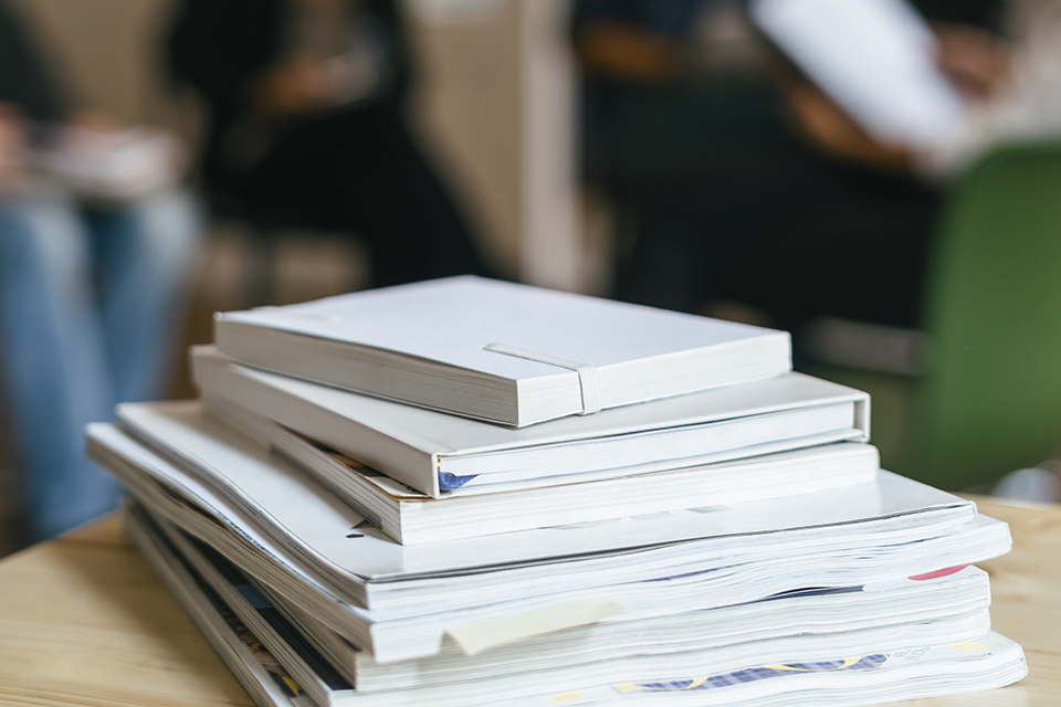 Pile of magazines and books on shelf, business peole talking in background